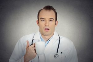 35516059 - portrait sleepy exhausted young funny doctor holding cup coffee tired busy day night shift trying to stay awake almost asleep isolated grey background. sleep deprivation. face expression emotion